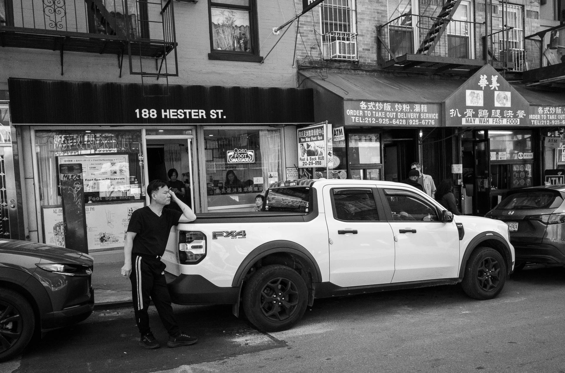Street + urban photography 145 Black-and-white street photograph of a man leaning against a white pickup truck outside storefronts on Hester Street in Manhattan’s Chinatown.