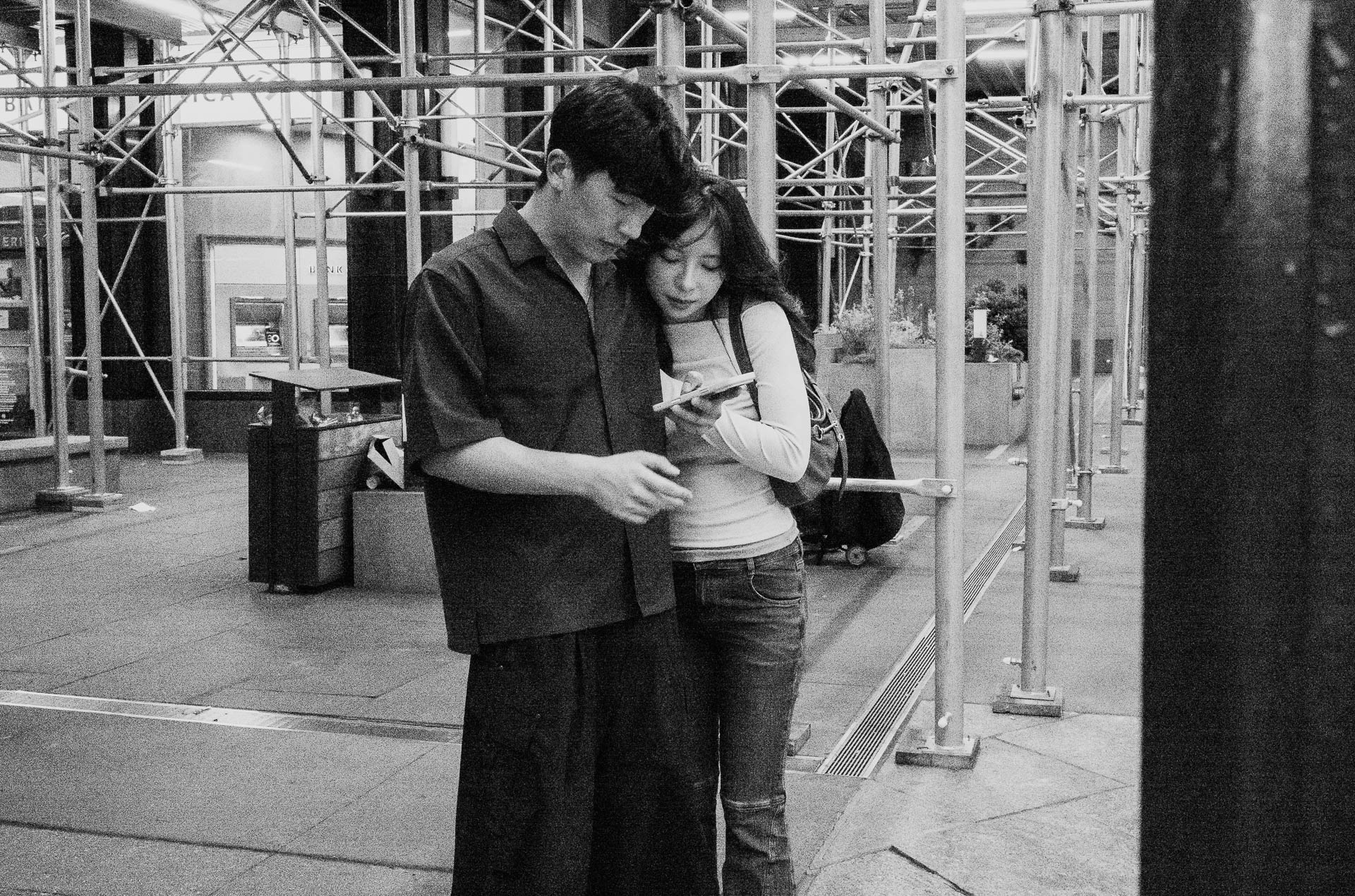 Street + urban photography 149 Black-and-white street photograph of a young couple standing closely together beneath sidewalk scaffolding while looking at a phone.