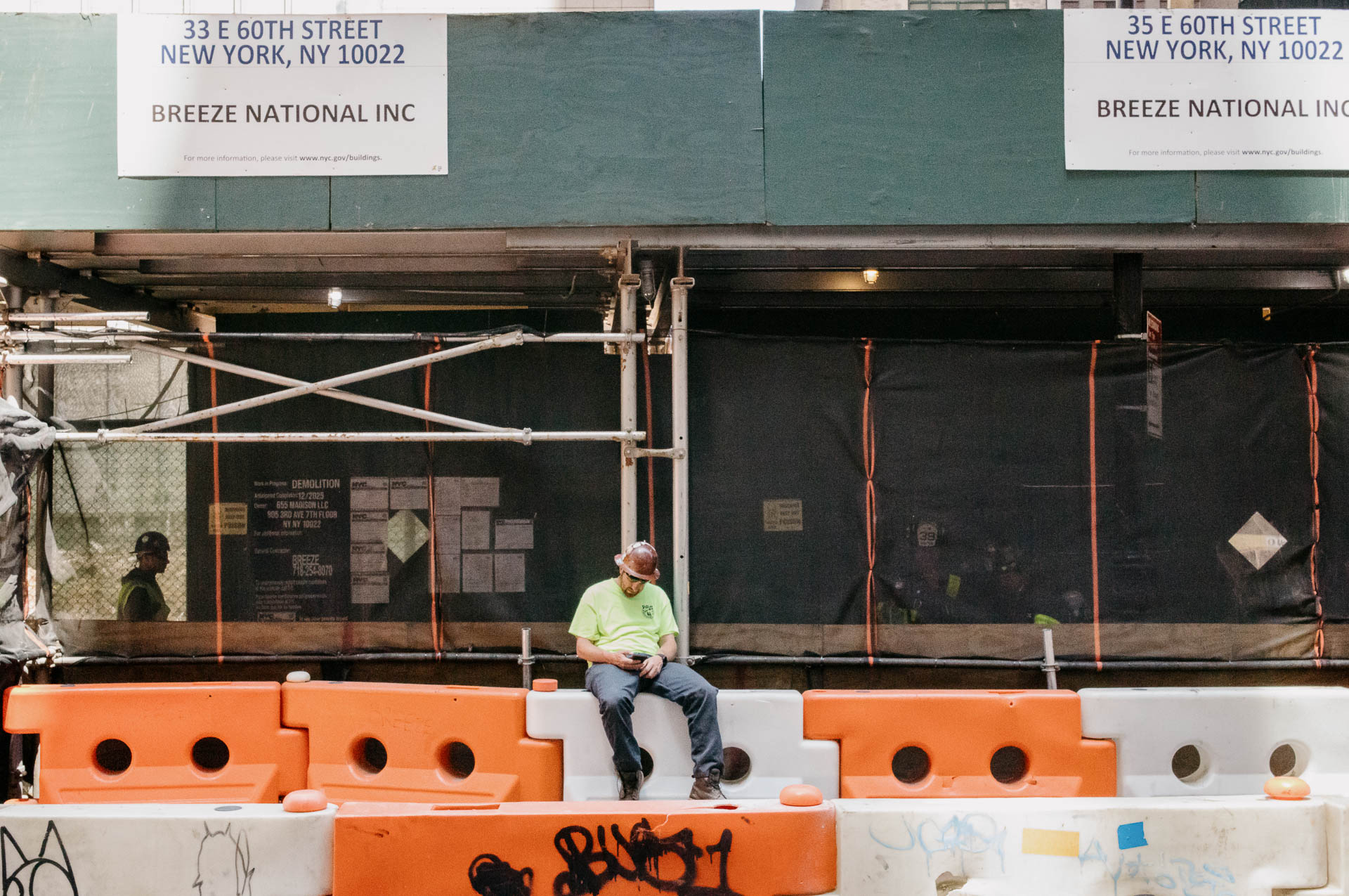 Street + urban photography 151 Color street photograph of a construction worker sitting alone on orange-and-white barriers beneath scaffolding on East 60th Street in Manhattan.