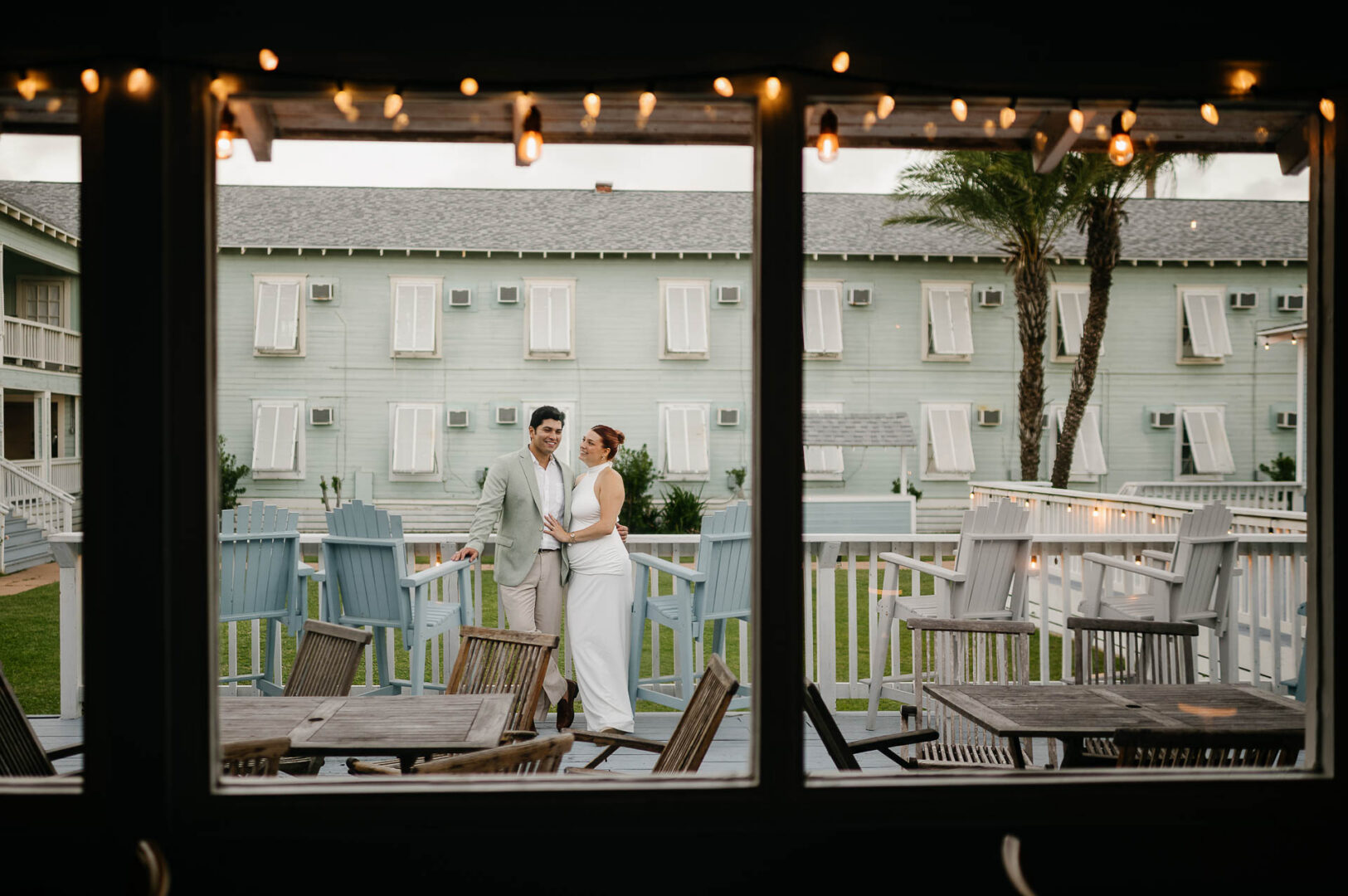 Chapel on the Dunes Wedding | Port Aransas, Texas | Mady+Roland 2 String lights hang beneath a covered patio at Roosevelt's in Port Aransas at dusk, with a couple walking in the distance past the hotel's balconied facade and palm trees.