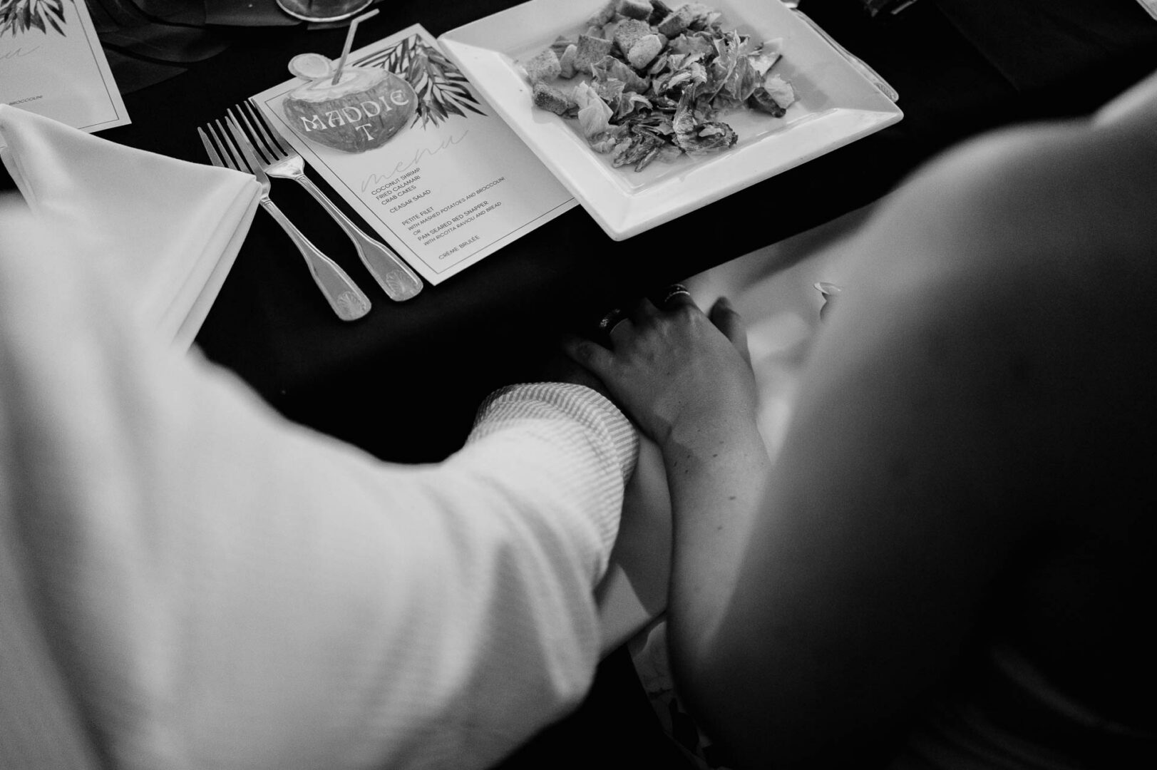 Chapel on the Dunes Wedding | Port Aransas, Texas | Mady+Roland 3 Black-and-white close-up over a guest's shoulder as they look at an illustrated rehearsal dinner program, background softly blurred.