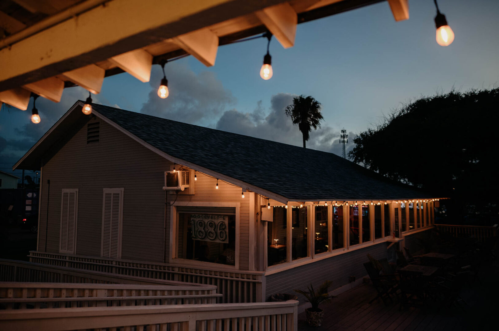Chapel on the Dunes Wedding | Port Aransas, Texas | Mady+Roland 4 Dusk shot of string lights strung along the eaves of a coastal building at Roosevelt's, a palm tree silhouetted against the fading blue sky.