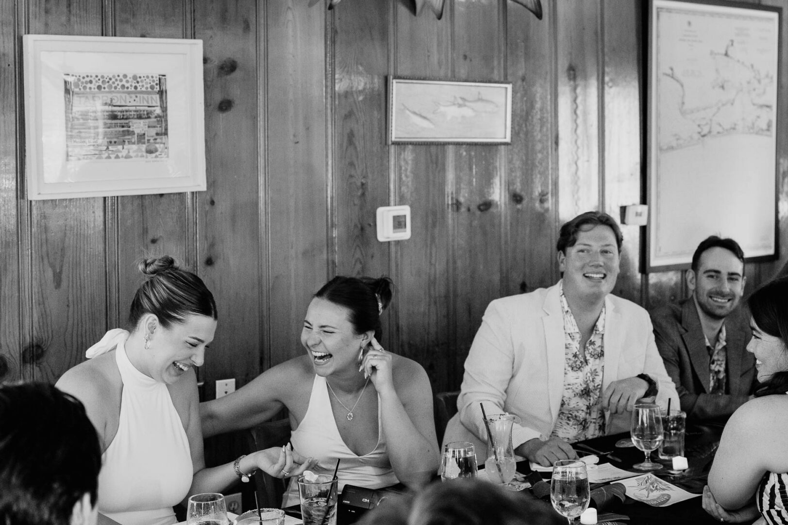 Chapel on the Dunes Wedding | Port Aransas, Texas | Mady+Roland 5 Black-and-white candid of two women laughing together at a wood-paneled table during the rehearsal dinner, framed art on the wall behind them.