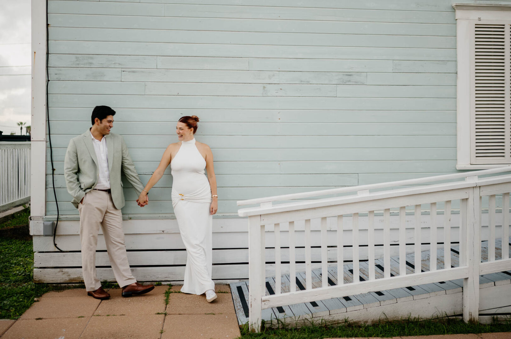 Chapel on the Dunes Wedding | Port Aransas, Texas | Mady+Roland 6 The couple walks hand-in-hand along the porch of a coastal cottage-style building at Roosevelt's, she in a white dress, he in a light suit.