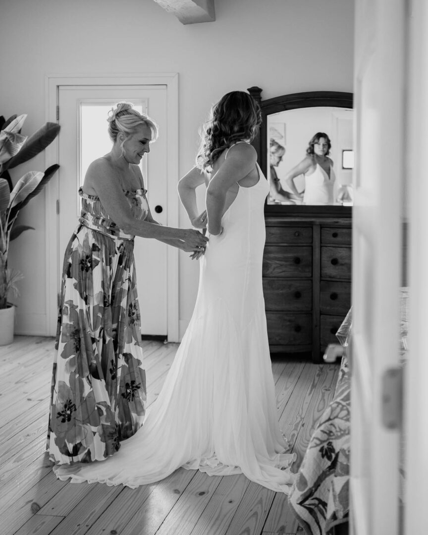 Chapel on the Dunes Wedding | Port Aransas, Texas | Mady+Roland 13 Black-and-white shot of a mother of the bride zipping up the back of the bride's gown in front of a mirror.