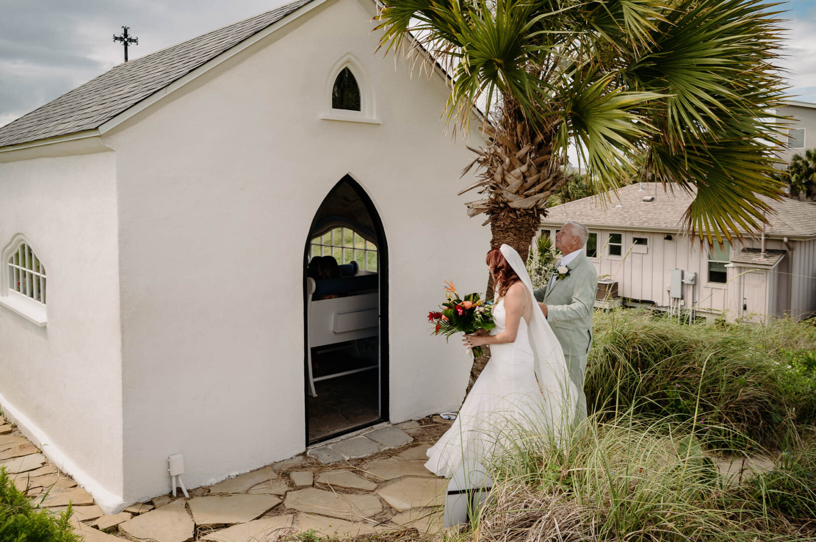 Chapel on the Dunes Wedding | Port Aransas, Texas | Mady+Roland 26 Exterior of the white chapel with an arched black door, framed by palm trees against a coastal sky.