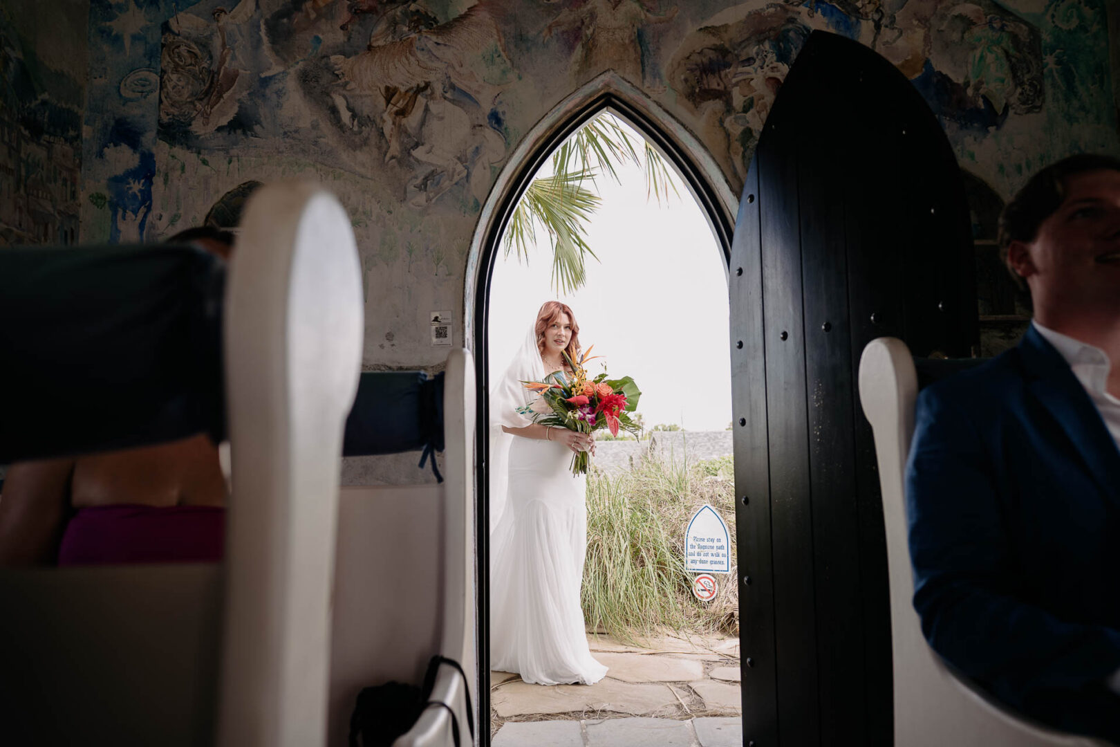 Chapel on the Dunes Wedding | Port Aransas, Texas | Mady+Roland 27 The bride stands in the chapel's arched doorway holding her bouquet, colorful stained glass visible inside.