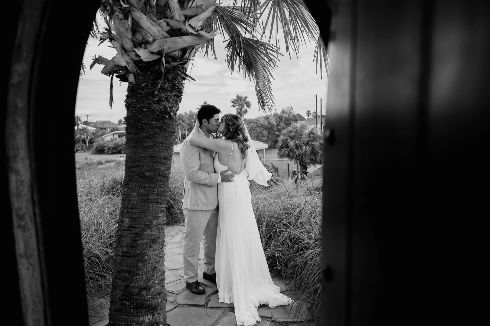 Chapel on the Dunes Wedding | Port Aransas, Texas | Mady+Roland 35 Black-and-white shot of the groom lifting and kissing the bride beneath palm trees.