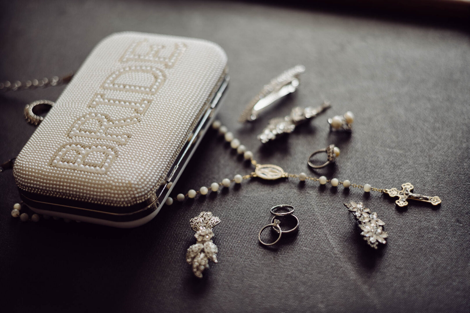 Tapatio Springs Hill Country Resort | Luana + Mack | Helotes 4 Pearl-studded "BRIDE" clutch lying on a dark surface beside a rosary, wedding bands, vintage hair clips and a pearl ring.