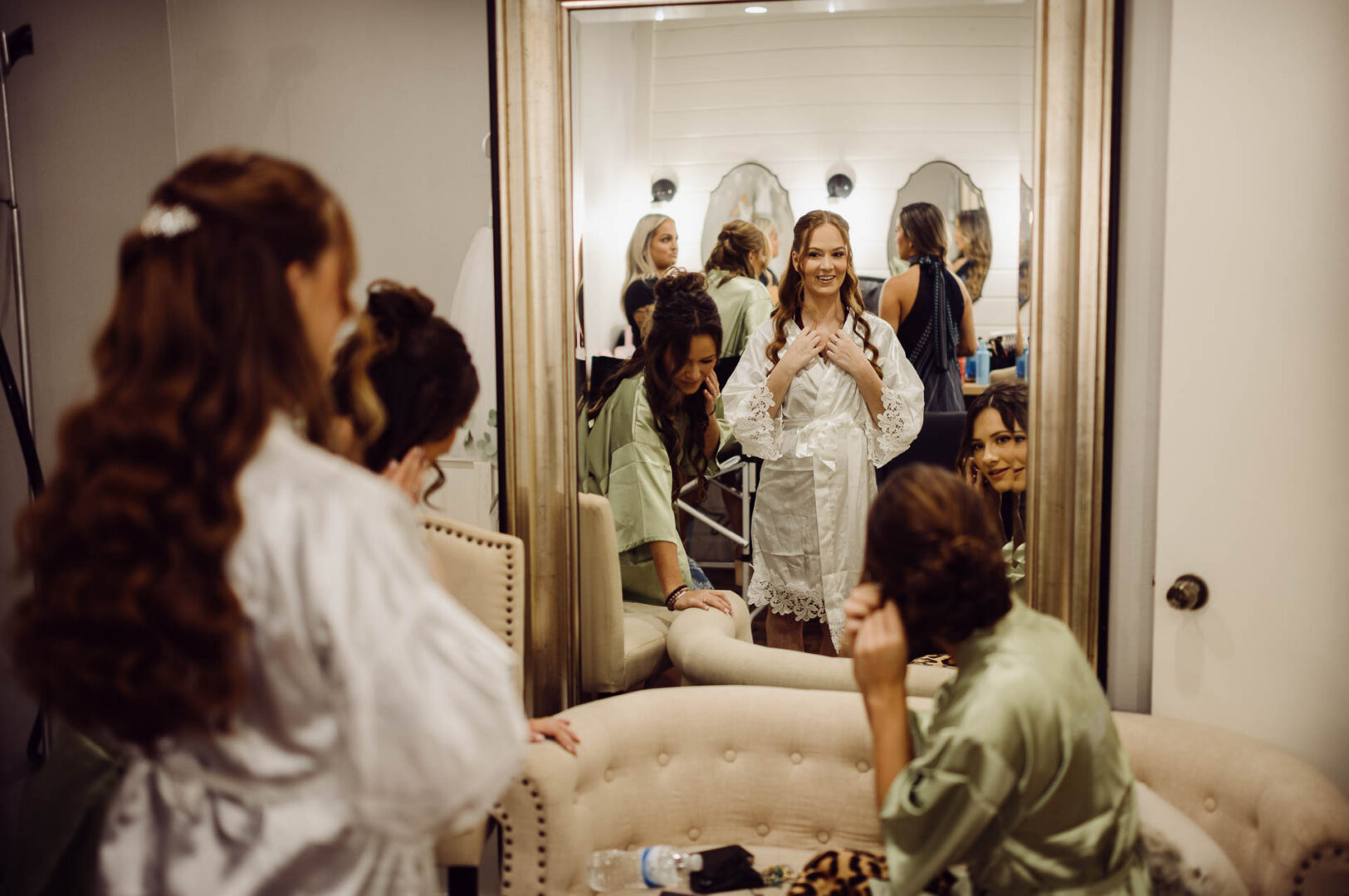 Tapatio Springs Hill Country Resort | Luana + Mack | Helotes 6 Bride in a white robe checks herself in a tall mirror in the getting-ready room, bridesmaids in sage green robes fixing their hair around her.