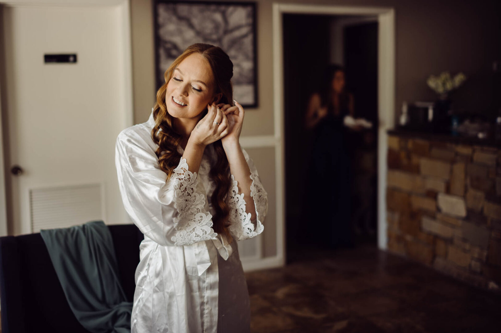 Tapatio Springs Hill Country Resort | Luana + Mack | Helotes 7 Bride in a lace-sleeved robe smiles with her eyes closed as she puts in an earring, a bridesmaid standing in the dark doorway behind her.