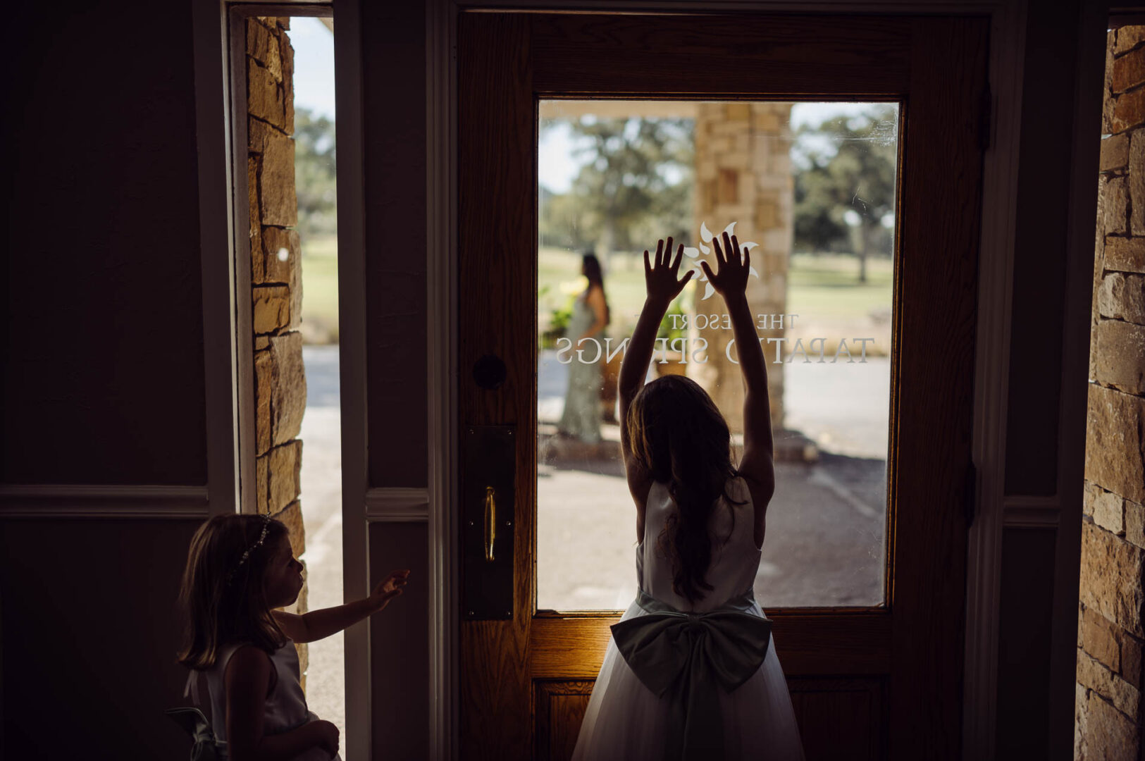 Tapatio Springs Hill Country Resort | Luana + Mack | Helotes 10 Silhouetted flower girl in a white dress with a big sage bow raises both hands against the glass of the resort door, while a smaller flower girl reaches toward her and a bridesmaid waits outside.