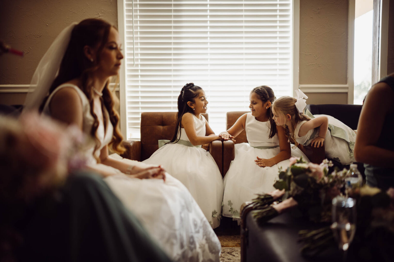 Tapatio Springs Hill Country Resort | Luana + Mack | Helotes 15 Three flower girls in white dresses laugh and talk on leather chairs by a bright window, with the bride blurred in the foreground.