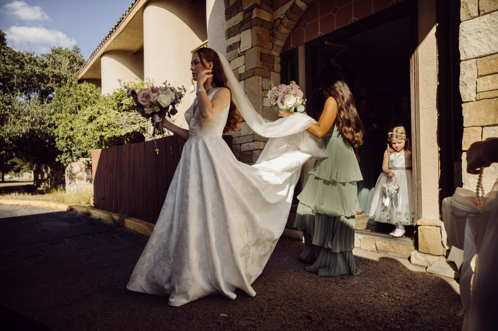 Tapatio Springs Hill Country Resort | Luana + Mack | Helotes 16 A bridesmaid in a sage green dress carries the bride's veil as she walks out into harsh sunlight, while a small flower girl waits in the doorway.