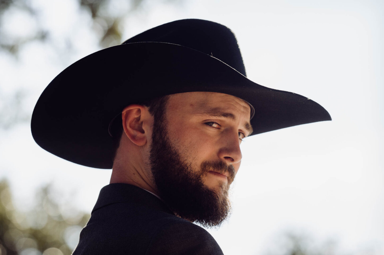 Tapatio Springs Hill Country Resort | Luana + Mack | Helotes 19 Close portrait of the groom in a black cowboy hat and full beard, glancing back over his shoulder in backlit sun.