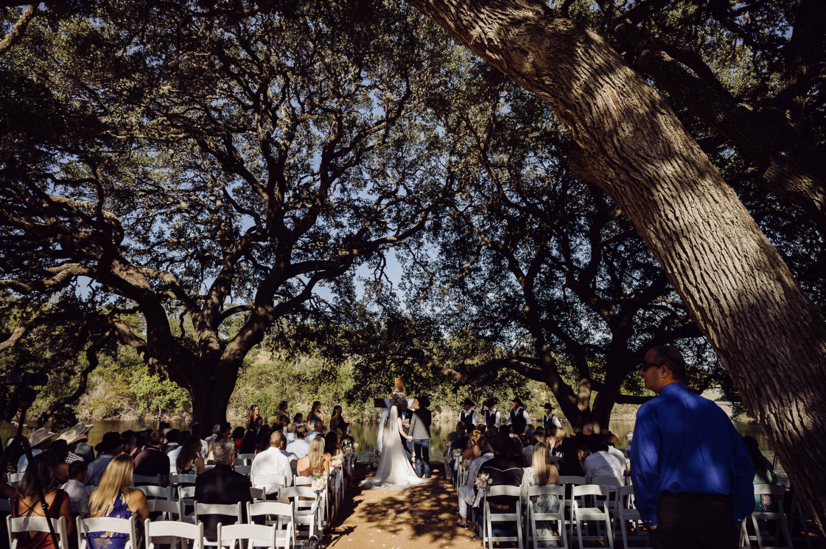 Tapatio Springs Hill Country Resort | Luana + Mack | Helotes 24 Wide view of the ceremony beneath a canopy of oak branches, guests seated on white chairs and the couple standing at a wooden cross by the water.