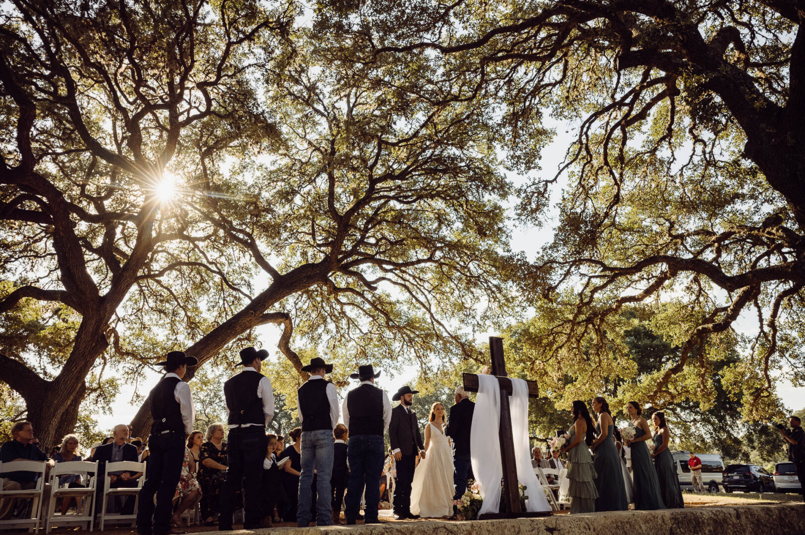 Tapatio Springs Hill Country Resort | Luana + Mack | Helotes 26 Low angle of the ceremony with sunlight bursting through the oaks, groomsmen and bridesmaids lined up on either side of the couple and a draped wooden cross.