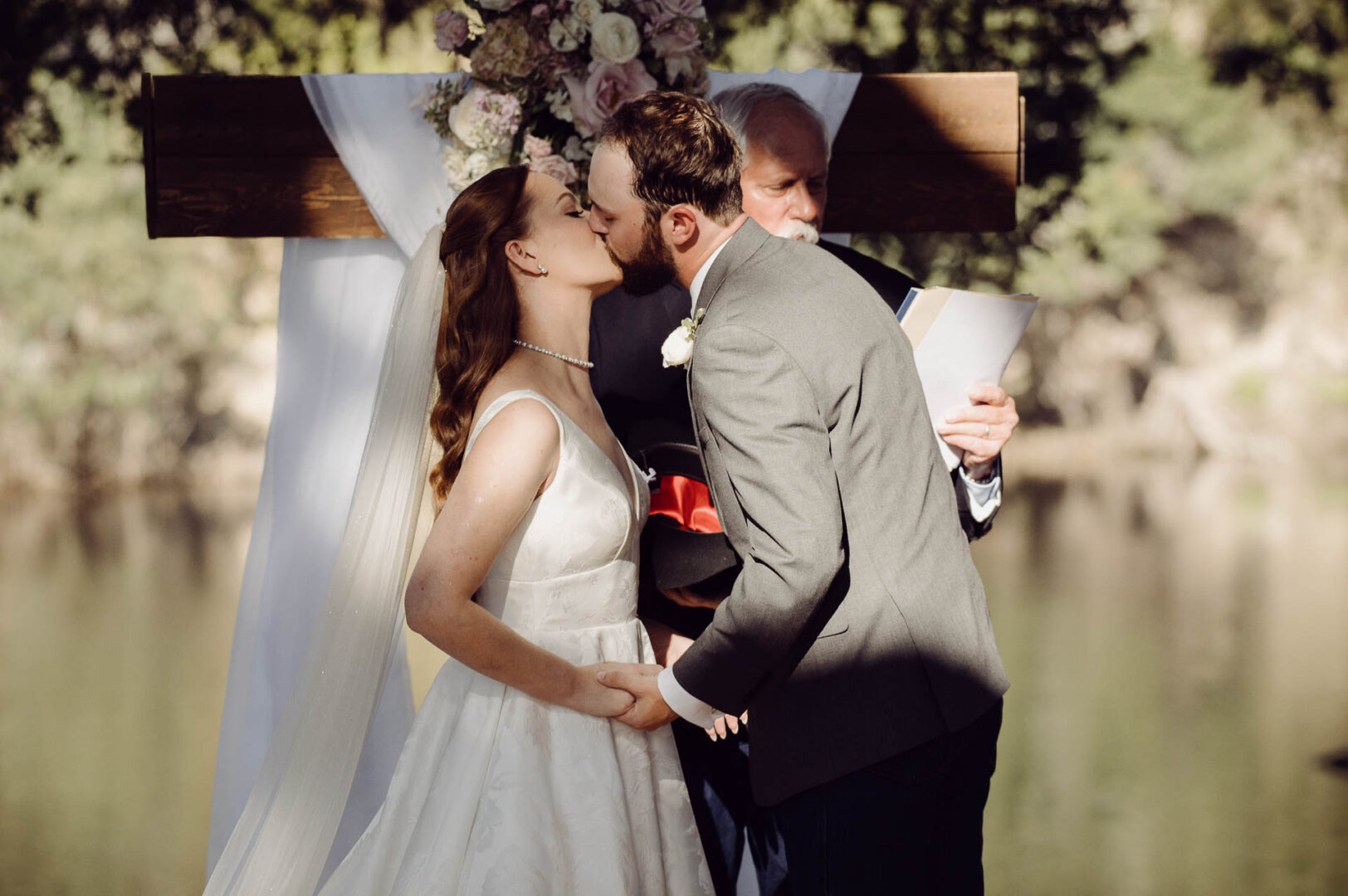 Tapatio Springs Hill Country Resort | Luana + Mack | Helotes 27 The couple share their first kiss in front of the wooden cross and pink and white florals, with the officiant and the pond behind.