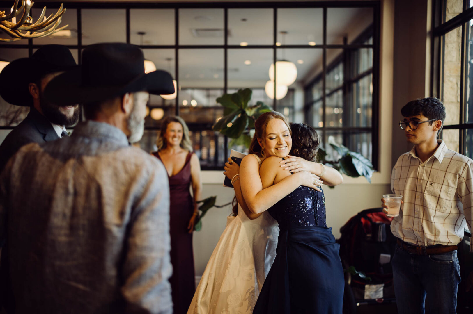 Tapatio Springs Hill Country Resort | Luana + Mack | Helotes 29 Bride hugs a woman in a navy beaded dress inside the restaurant, smiling with her eyes closed while guests look on.